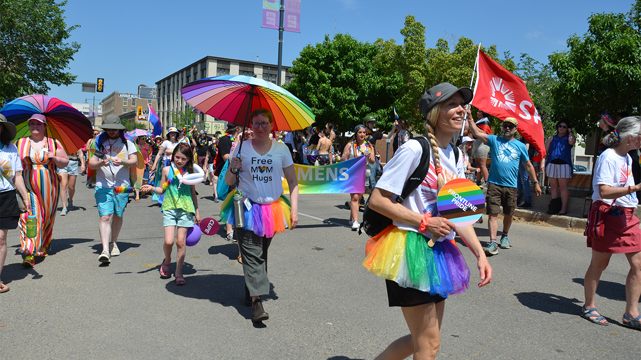 A group of library employees wearing colourful clothes and holding rainbow umbrellas walk in the Pride Parade in Saskatoon. An employee wearing a blue shirt is holding a red and white SPL flag. 